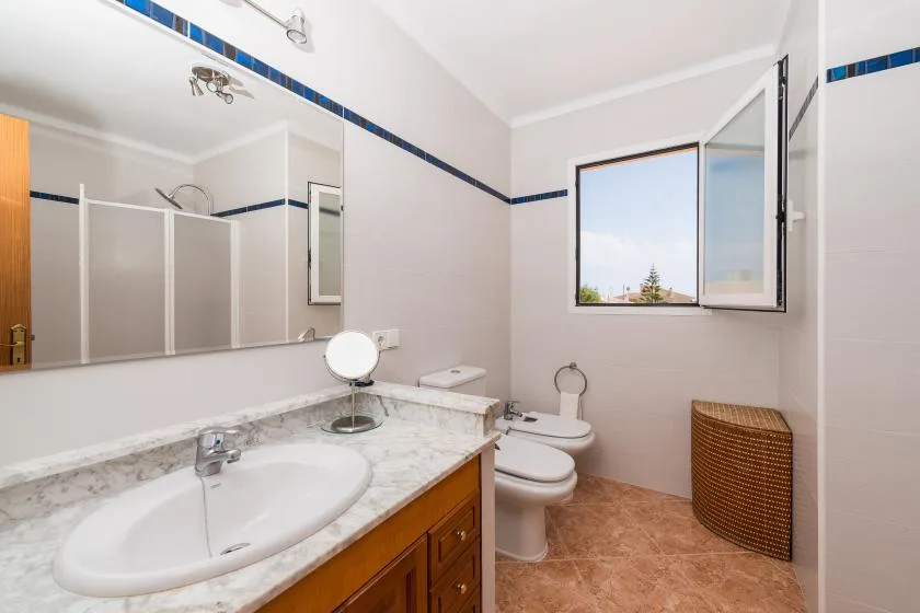 A bright bathroom with a long vanity, toilet, and open window.