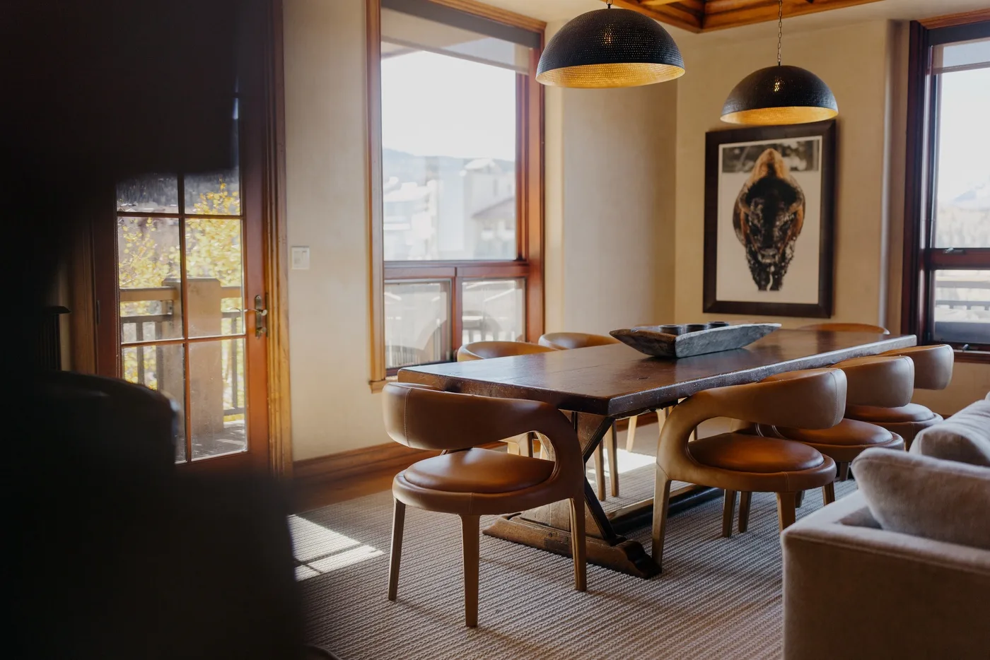 A dining area with a long table, seating, and large windows.