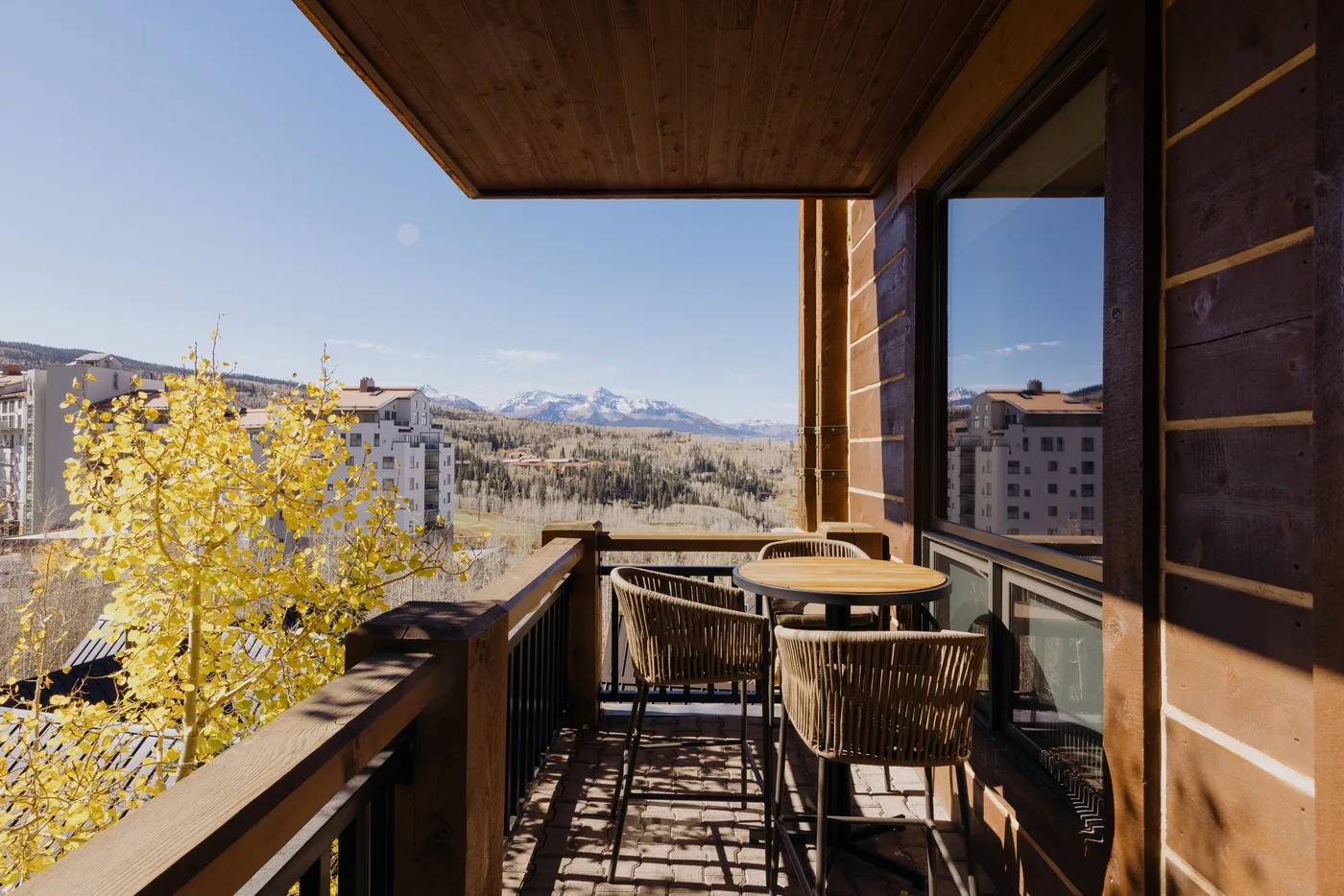A balcony with outdoor seating overlooks nearby buildings and distant mountains.