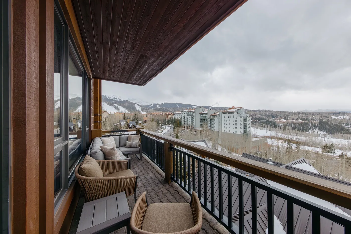 A covered balcony with outdoor seating overlooks buildings and snow-dusted hills.
