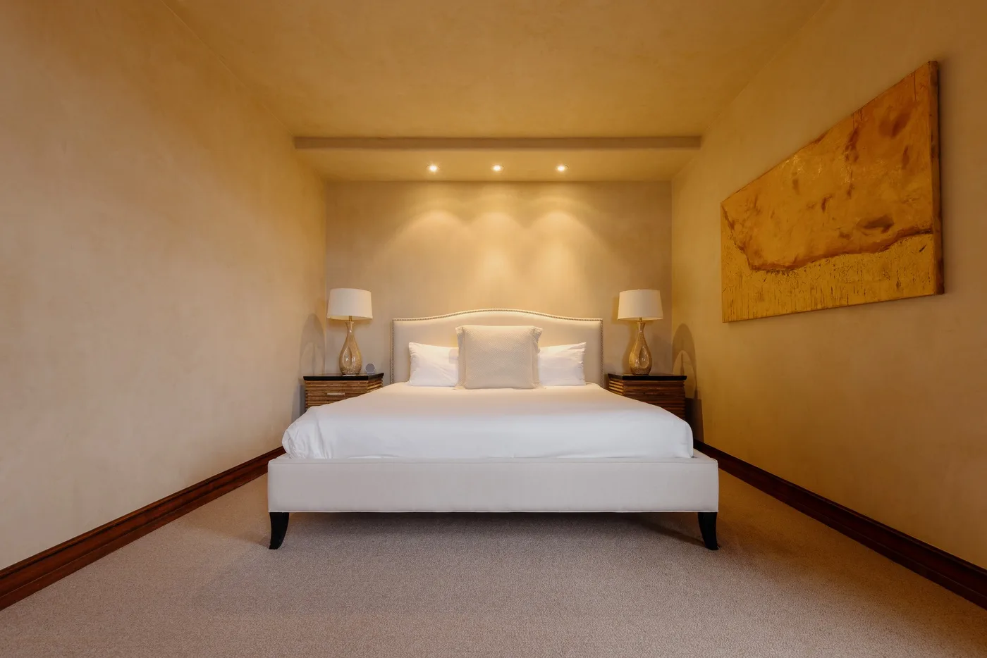 A bedroom with a bed, bedside tables, and warm recessed lighting.
