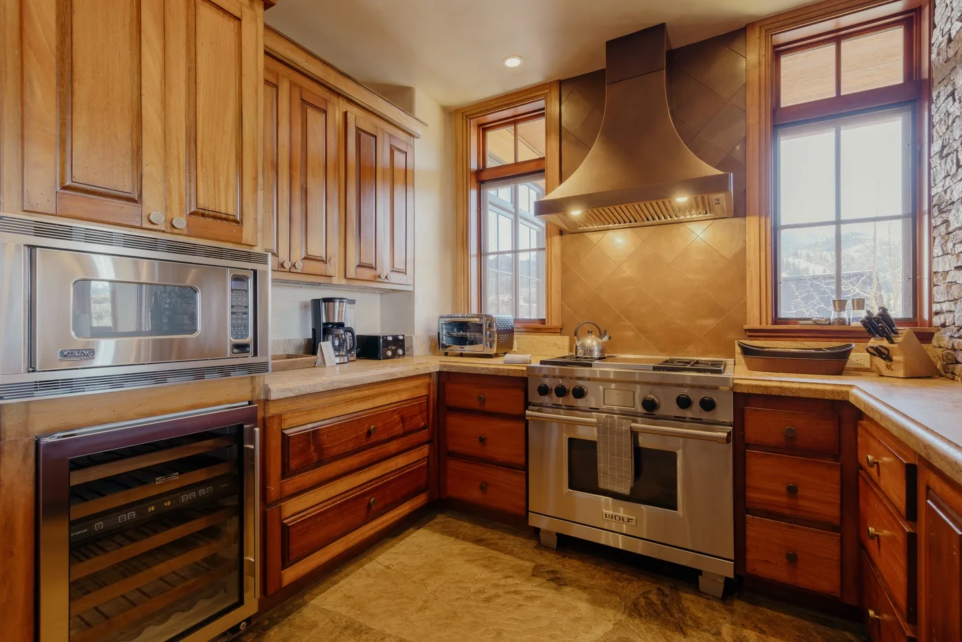 A warm-toned kitchen with wooden cabinets and a large range beneath a metal hood.