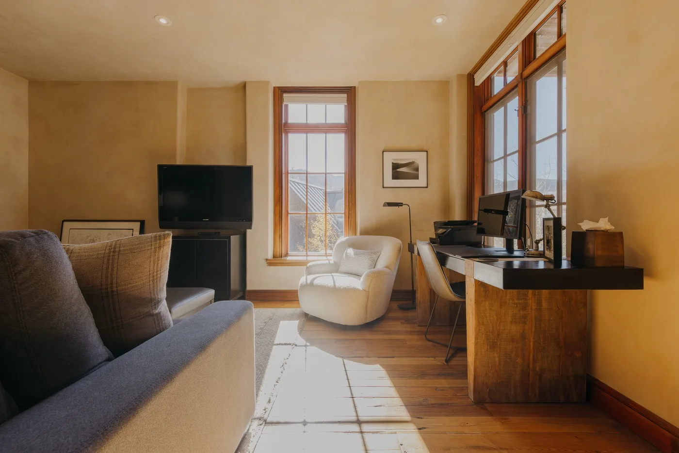 A sunlit living room with seating, a television, and a desk by the windows.