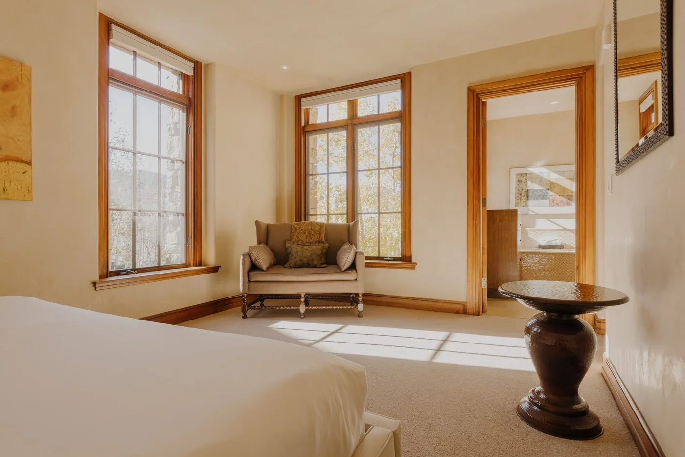 A sunlit bedroom with a bed, a seating area, and tall windows.