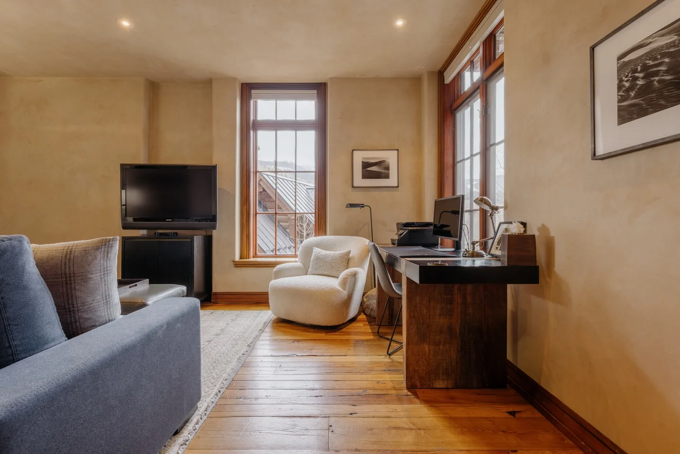 A living room with a television, seating, and a desk beside tall windows.