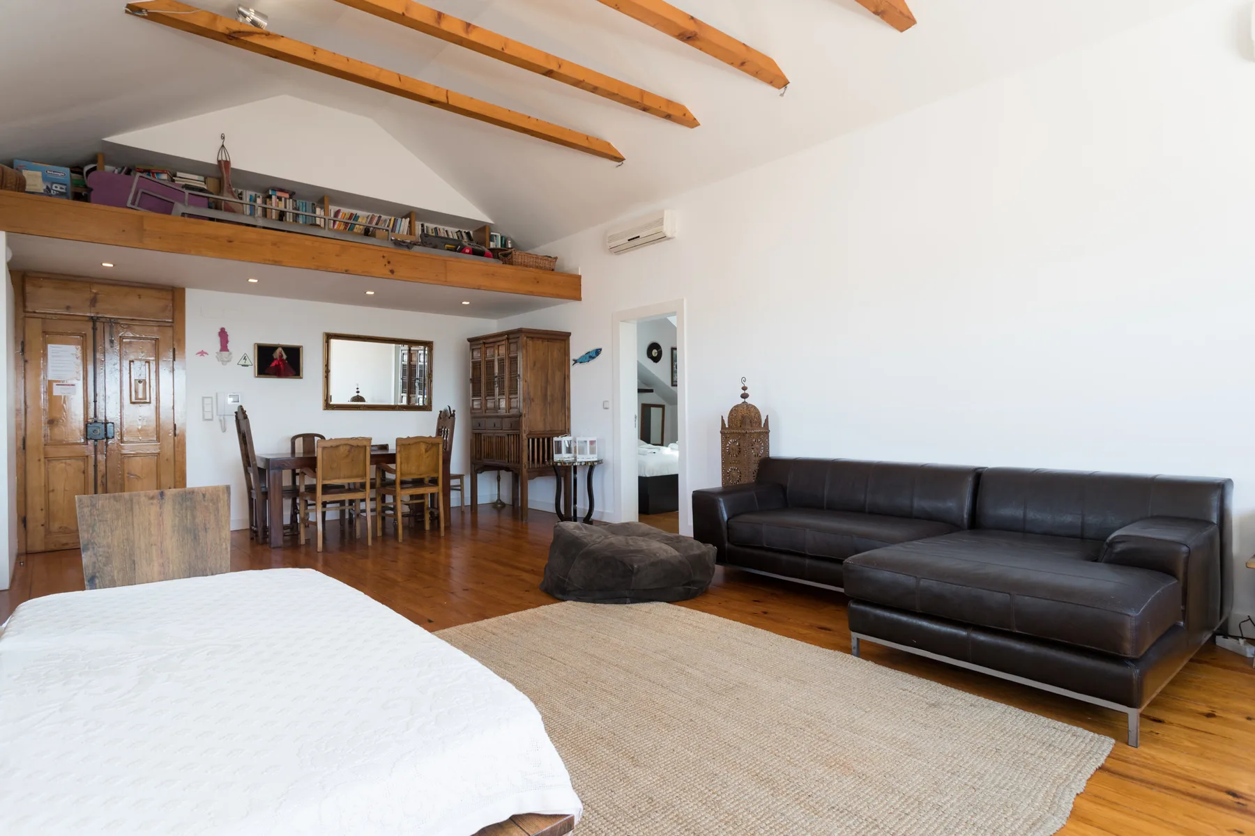 Una habitación amplia combina una zona para dormir, una mesa de comedor y asientos oscuros bajo vigas expuestas.