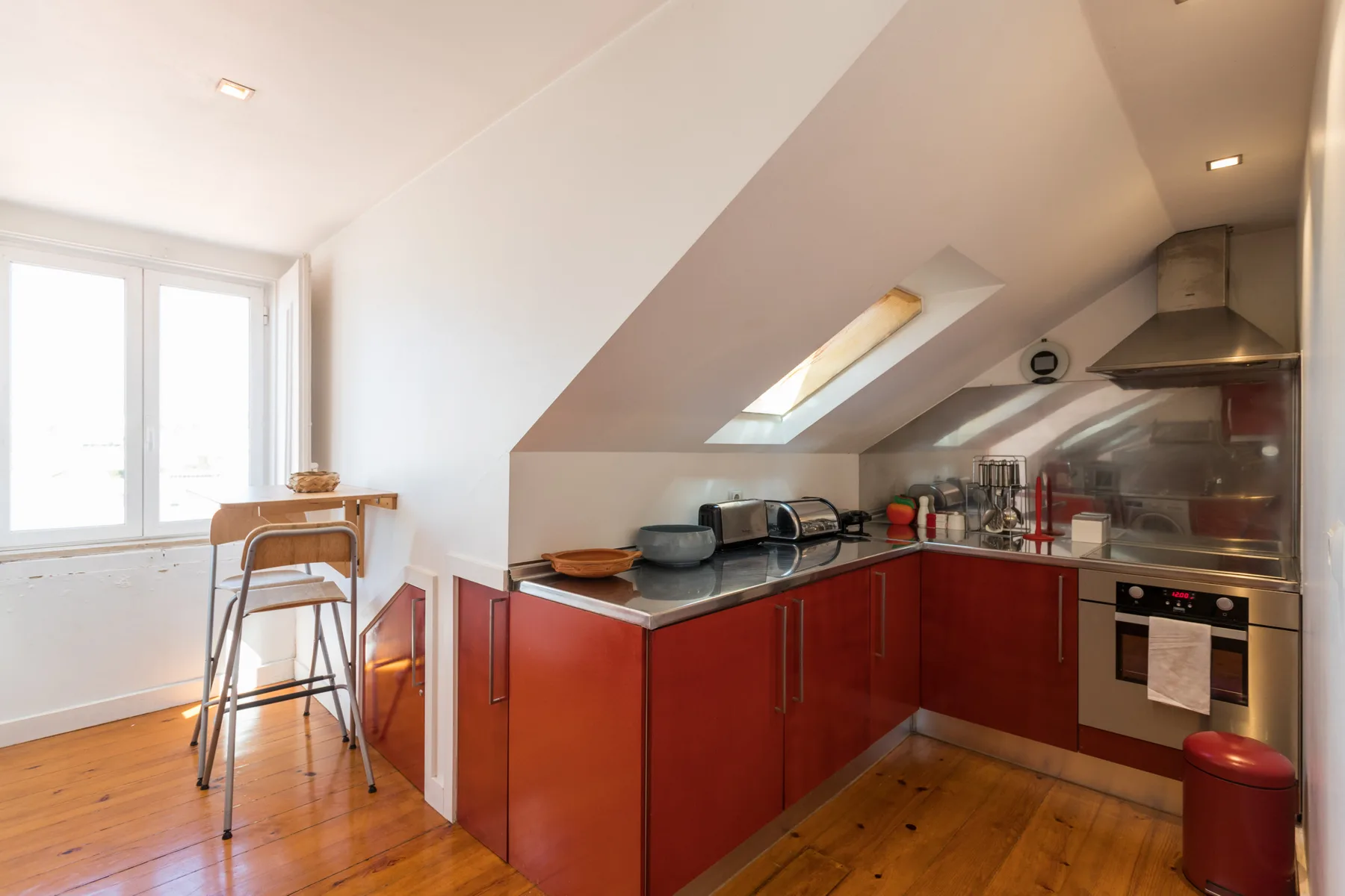 Una cocina compacta con gabinetes rojos está bajo un techo inclinado junto a una ventana luminosa.