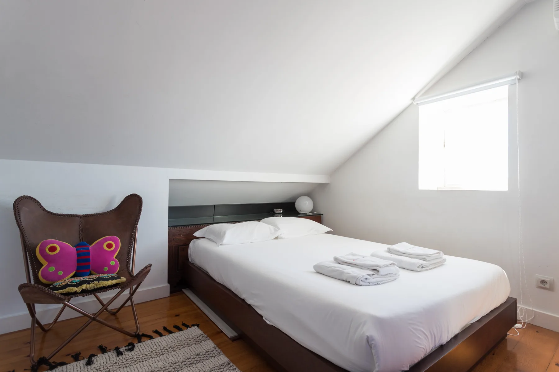 Un dormitorio luminoso con una cama y un cojín con estampado de mariposa en la silla.