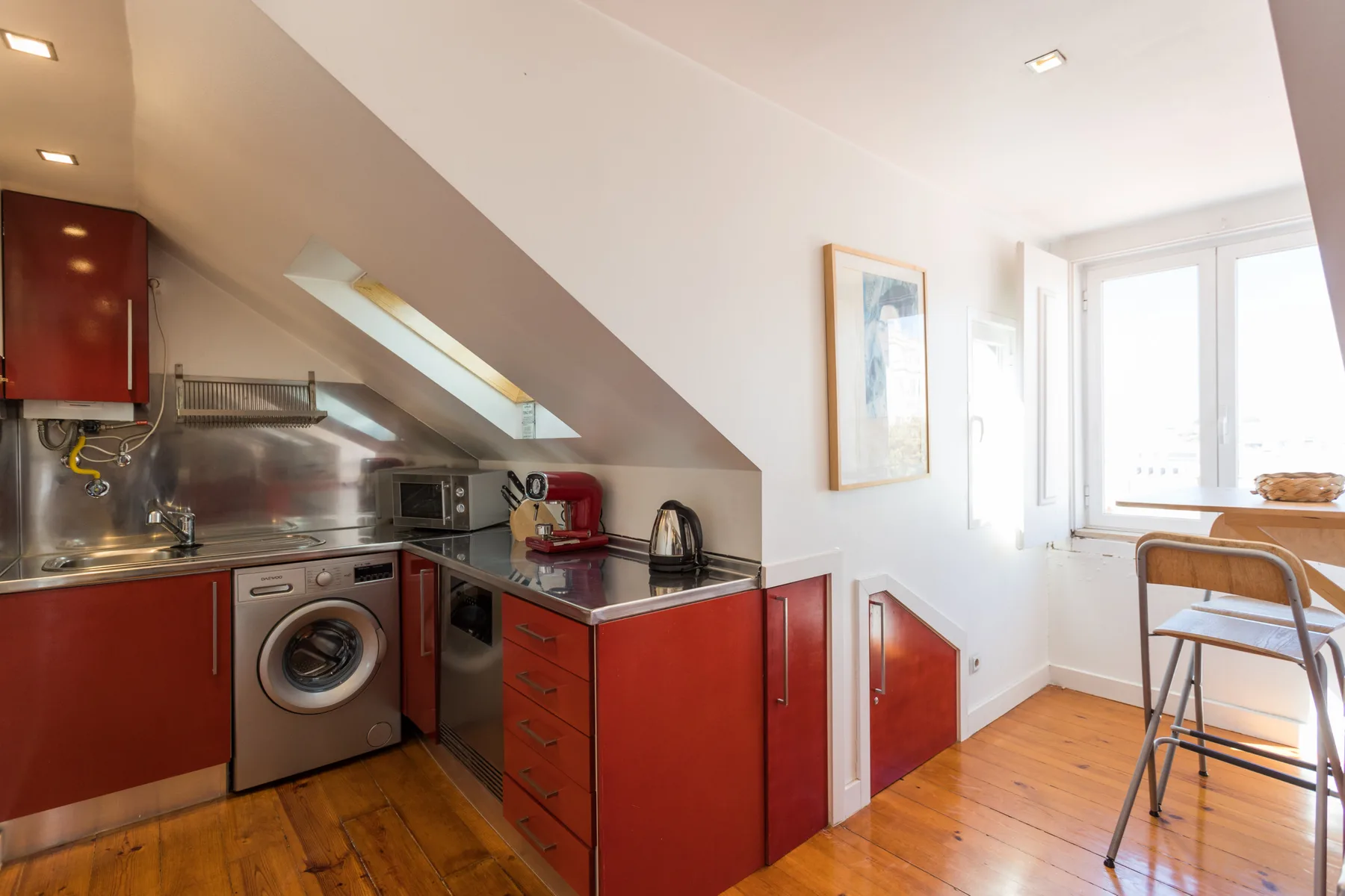 Una cocina compacta con gabinetes rojos y una lavadora bajo un techo inclinado.