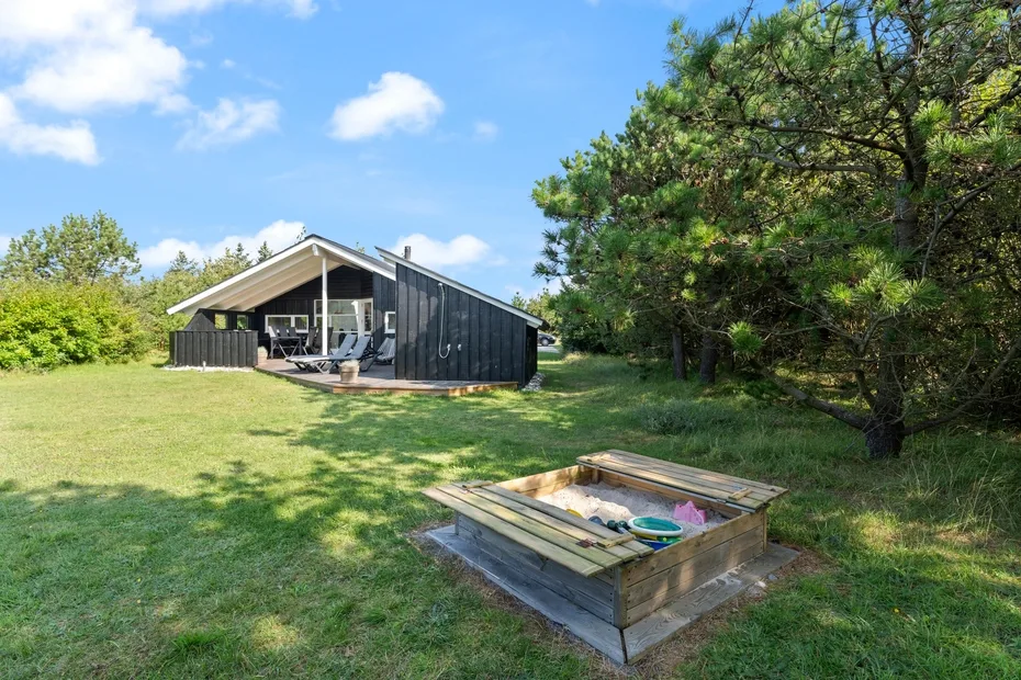A modern cabin sits beside a grassy yard with a wooden sandbox in the foreground.