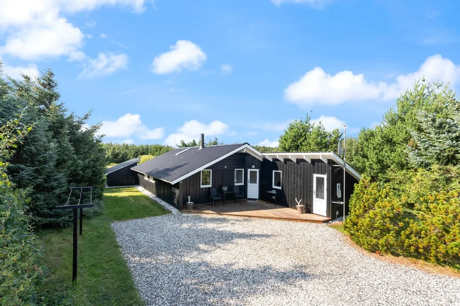 A dark wooden house stands beside a gravel driveway and green lawn.