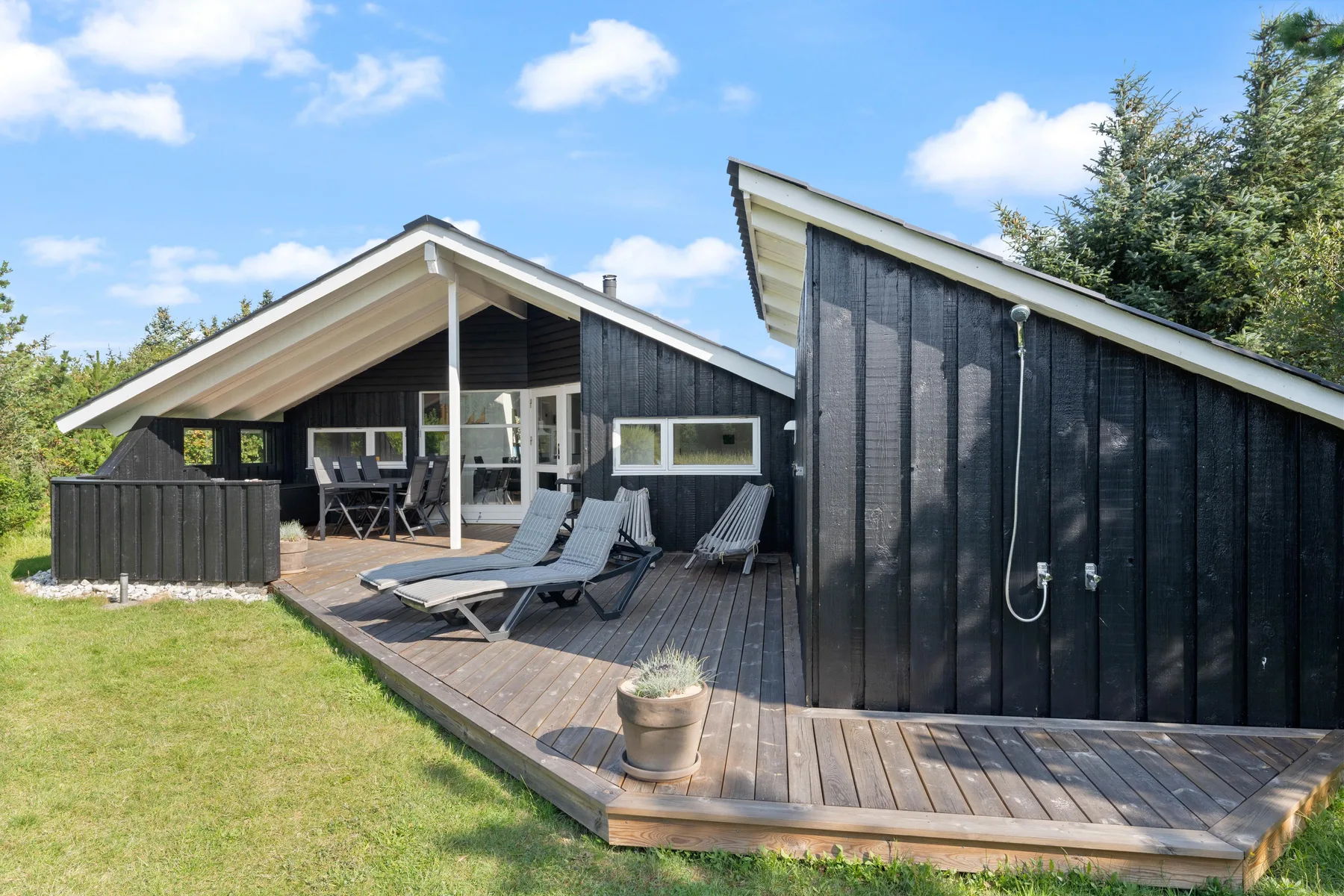 A dark-sided house with a raised wooden deck, lounge seating, and an outdoor shower.