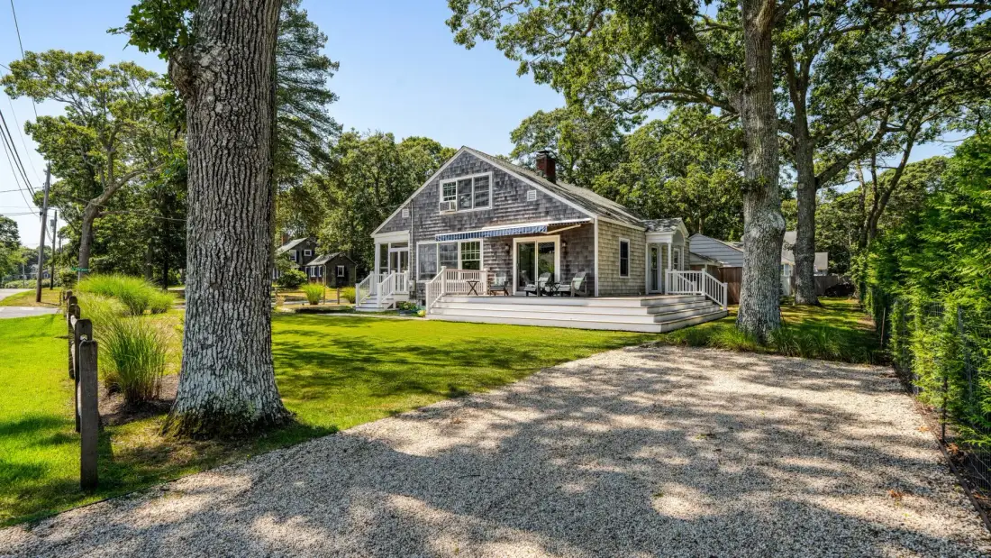A house stands beyond a gravel driveway, surrounded by trees and lawn.