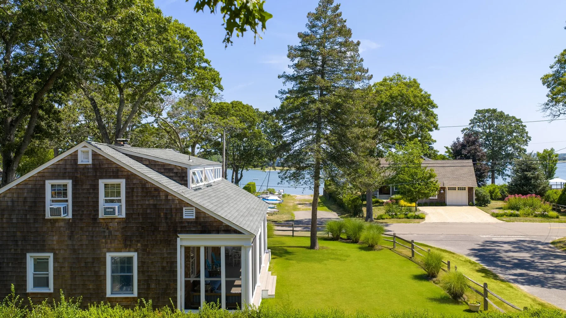 A lakeside house sits beside a green lawn, trees, and a paved road.