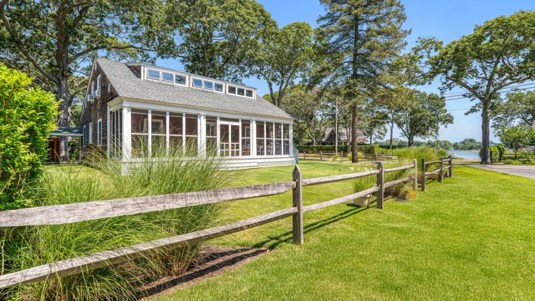 A house with a long glass-enclosed porch stands beside a lawn and wooden fence.