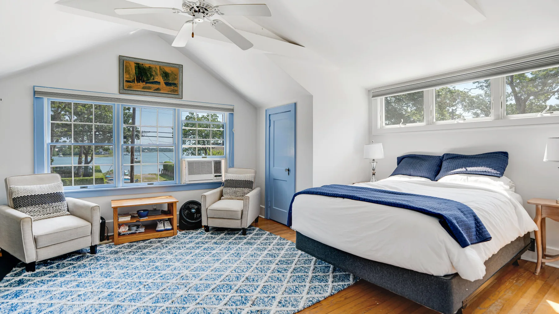 A bedroom with a large bed, two armchairs, and windows overlooking the water.
