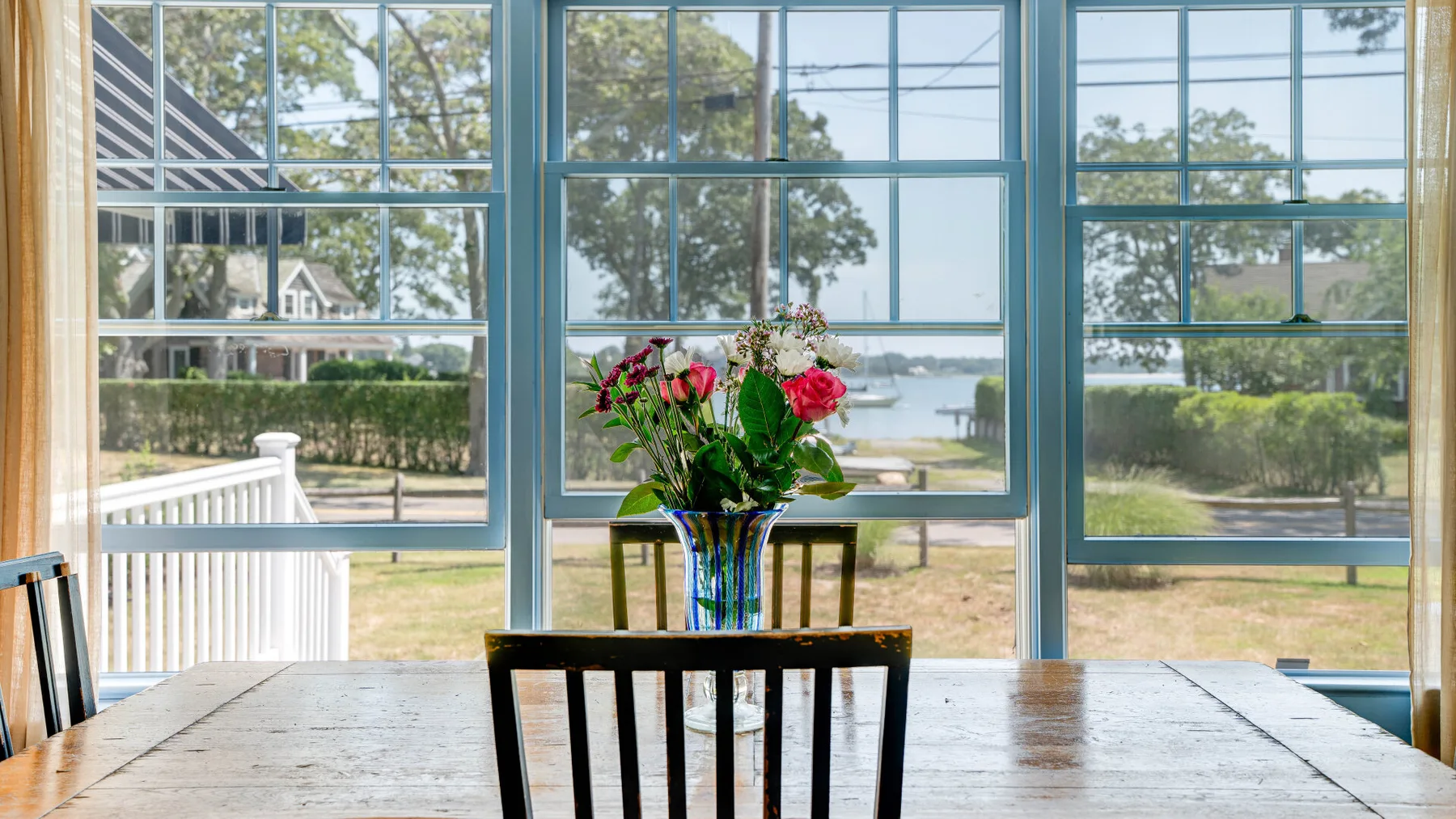 A dining area with a wooden table and flowers beside large windows overlooking a yard.