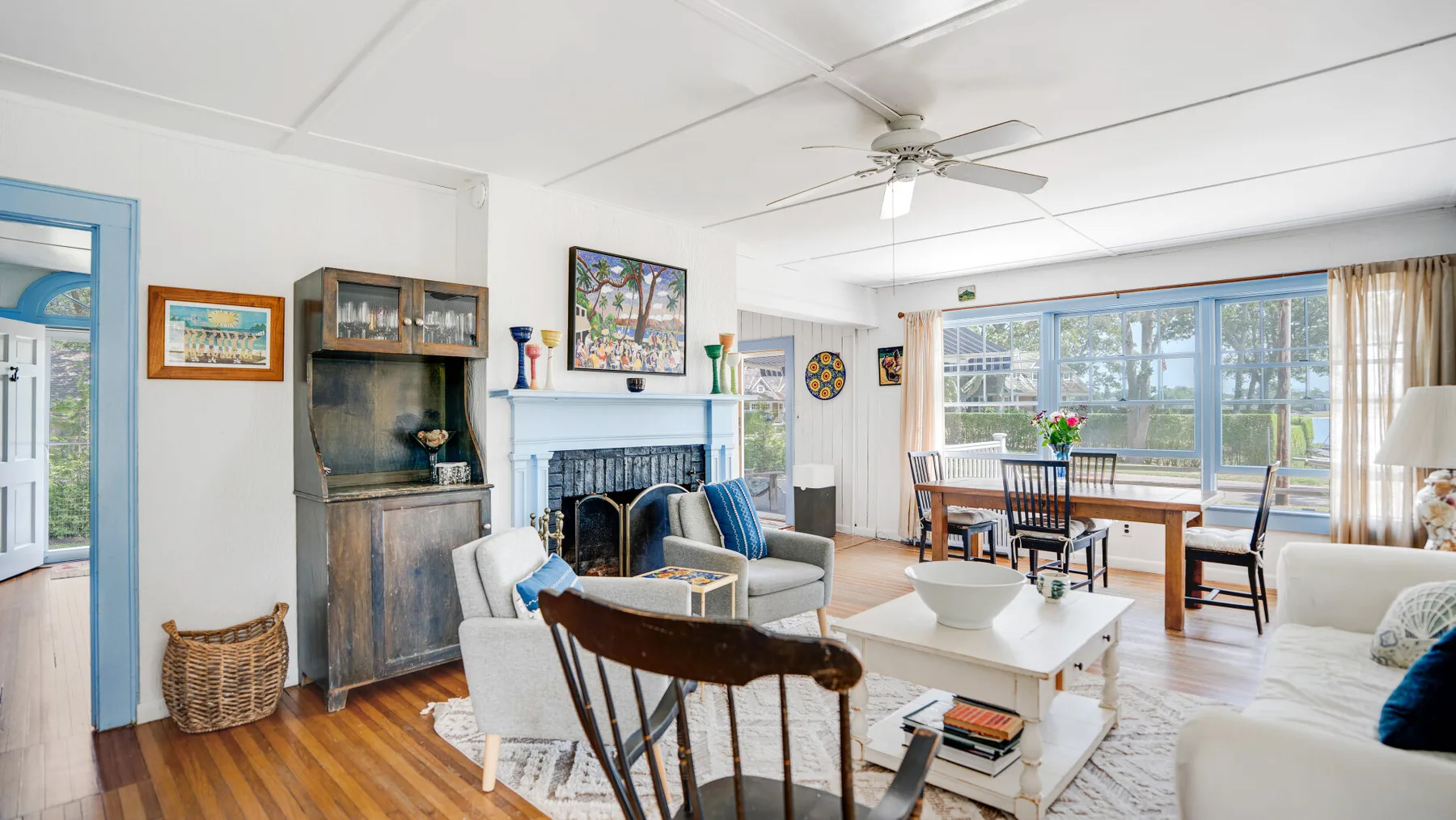 A bright living and dining room with seating, a fireplace, and wide windows.