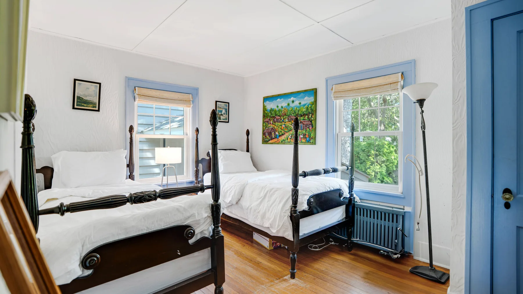 A bright bedroom with two beds, blue-trimmed windows, and framed artwork.