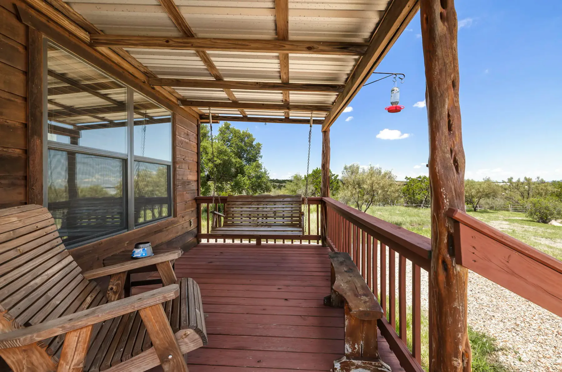 Wooden porch with rocking chairs, a hanging bird feeder, and a swing overlooking a grassy landscape under a blue sky.