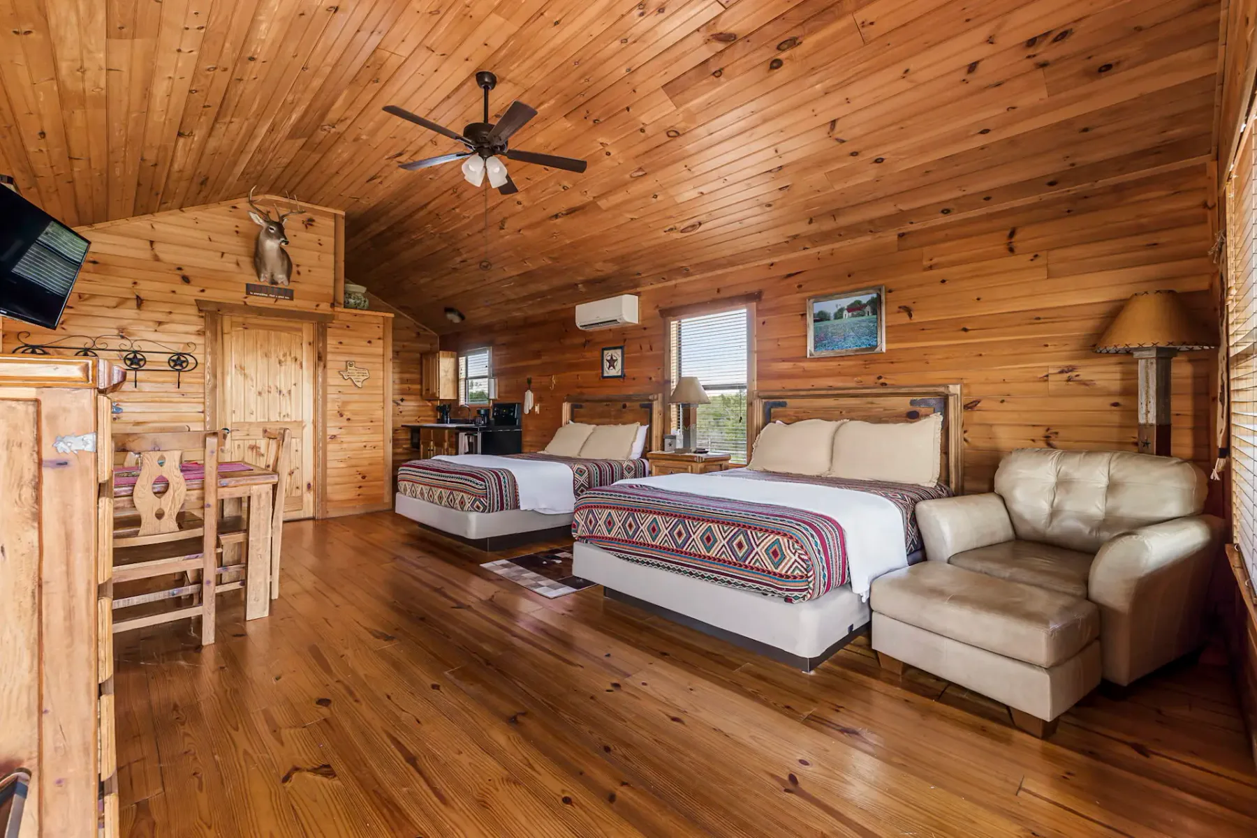 Cozy wooden cabin interior with two beds, a sofa, a small table, and a ceiling fan. Natural light enters through a window.