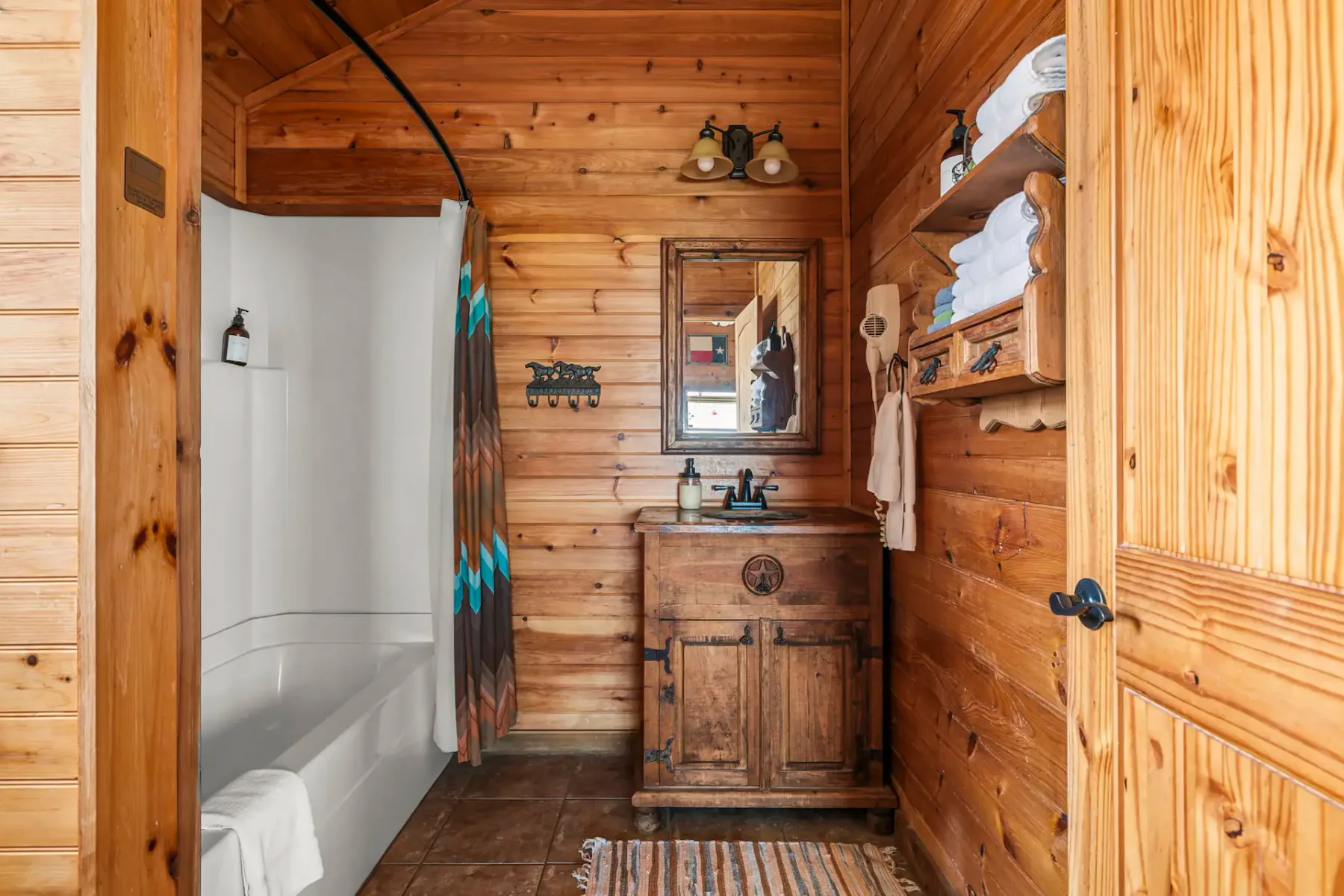 Rustic bathroom with wooden walls, a white bathtub with a shower curtain, a wooden vanity, mirror, and shelves holding towels. Rug on the tile floor.