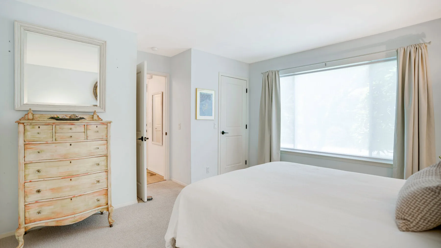 Un dormitorio luminoso con cama, cómoda y una gran ventana con cortinas.