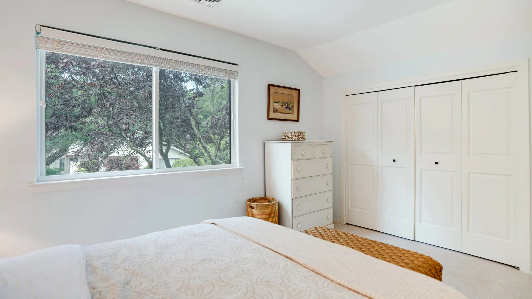 Un dormitorio luminoso con una cama, una cómoda y una ventana grande con vista a los árboles.
