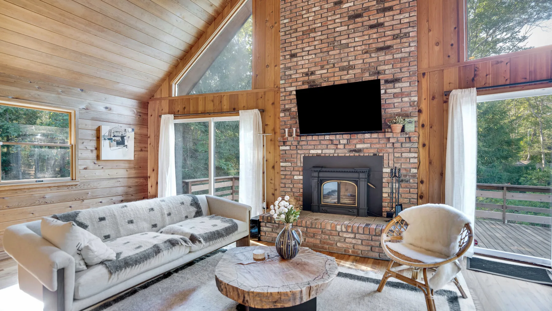 A vaulted living room features a brick fireplace, television, and seating by large windows.