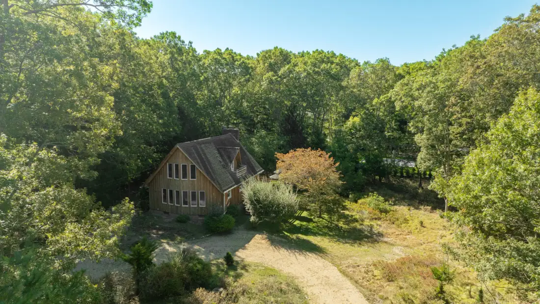 A wooden house sits beside a sunlit clearing surrounded by trees.