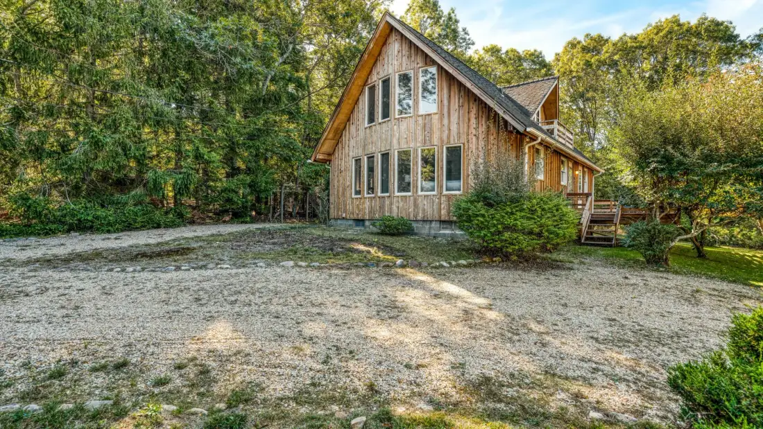 A wooden house stands beside a broad gravel clearing surrounded by trees.
