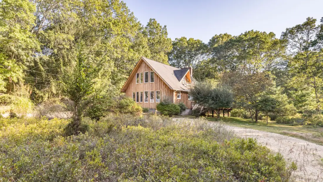 A wooden house sits among trees beside a gravel driveway.