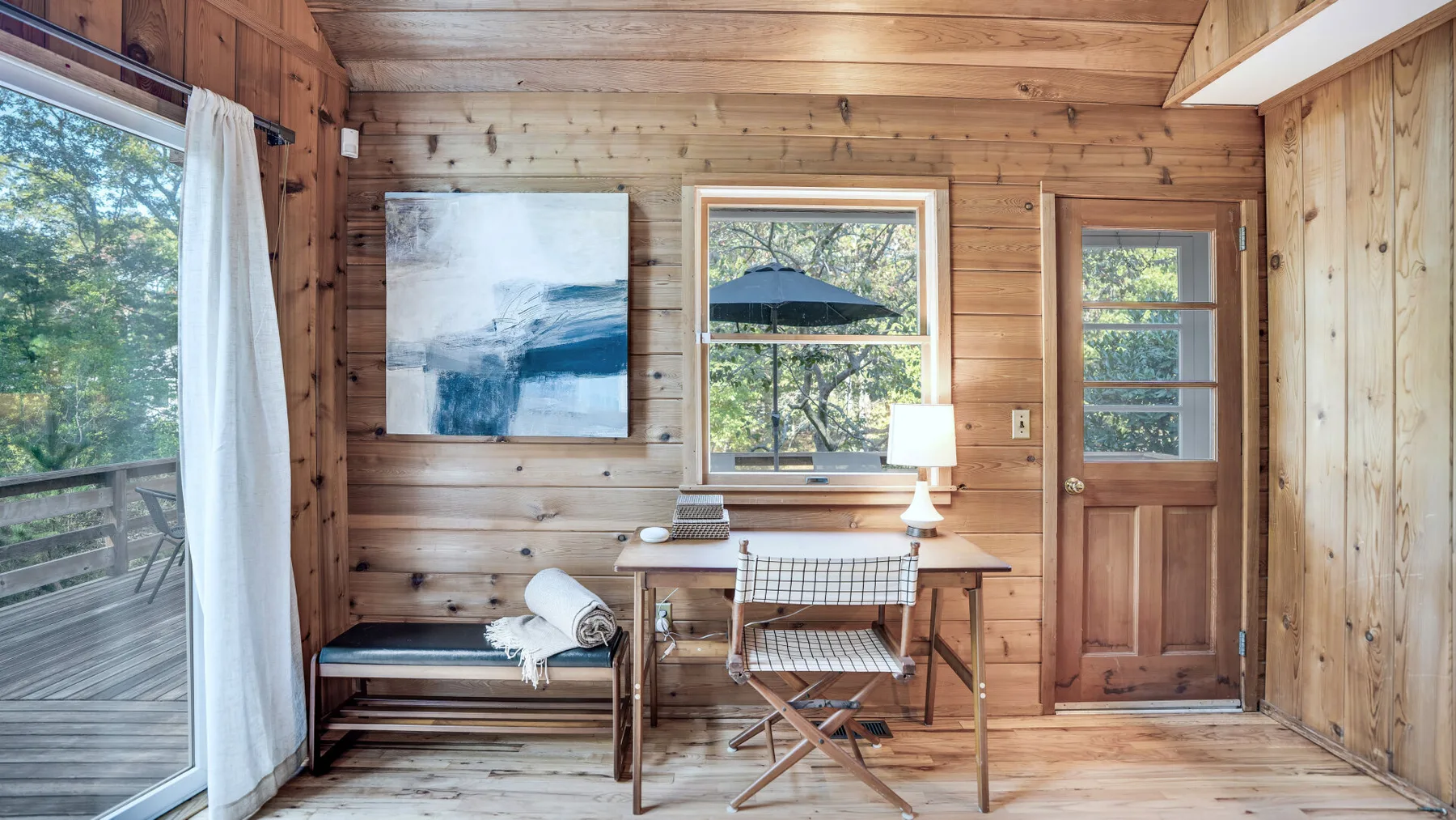 A wood-paneled room with a desk, chair, and windows overlooking greenery.