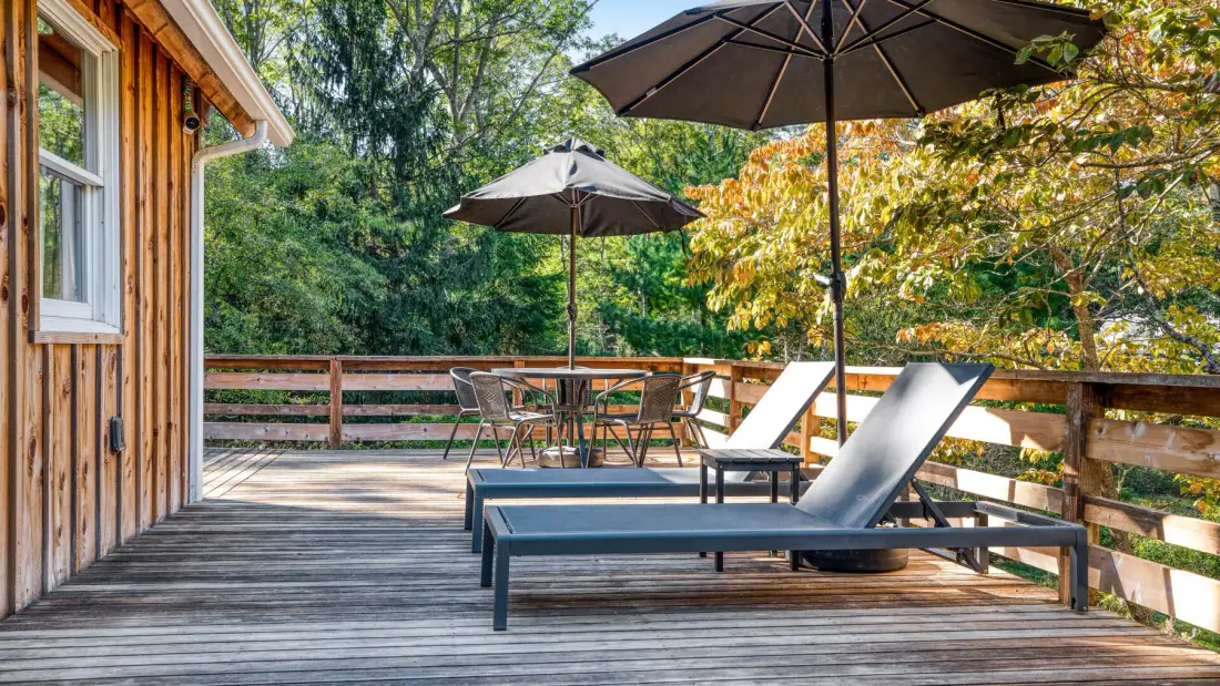 A wooden deck has lounge seating, tables, and umbrellas beside a tree-lined railing.