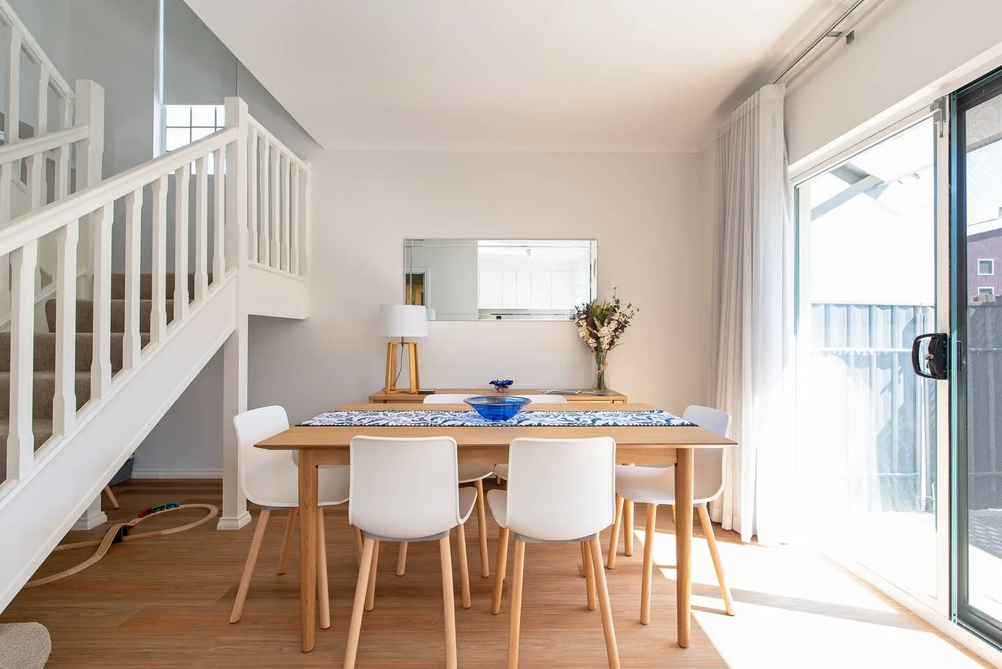 Una mesa de comedor con asientos blancos está bajo una escalera, junto a puertas corredizas de vidrio.