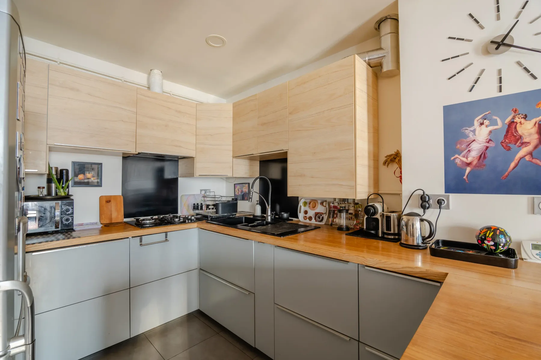 A modern kitchen with L-shaped counters, light wood cabinets, and dark appliances.