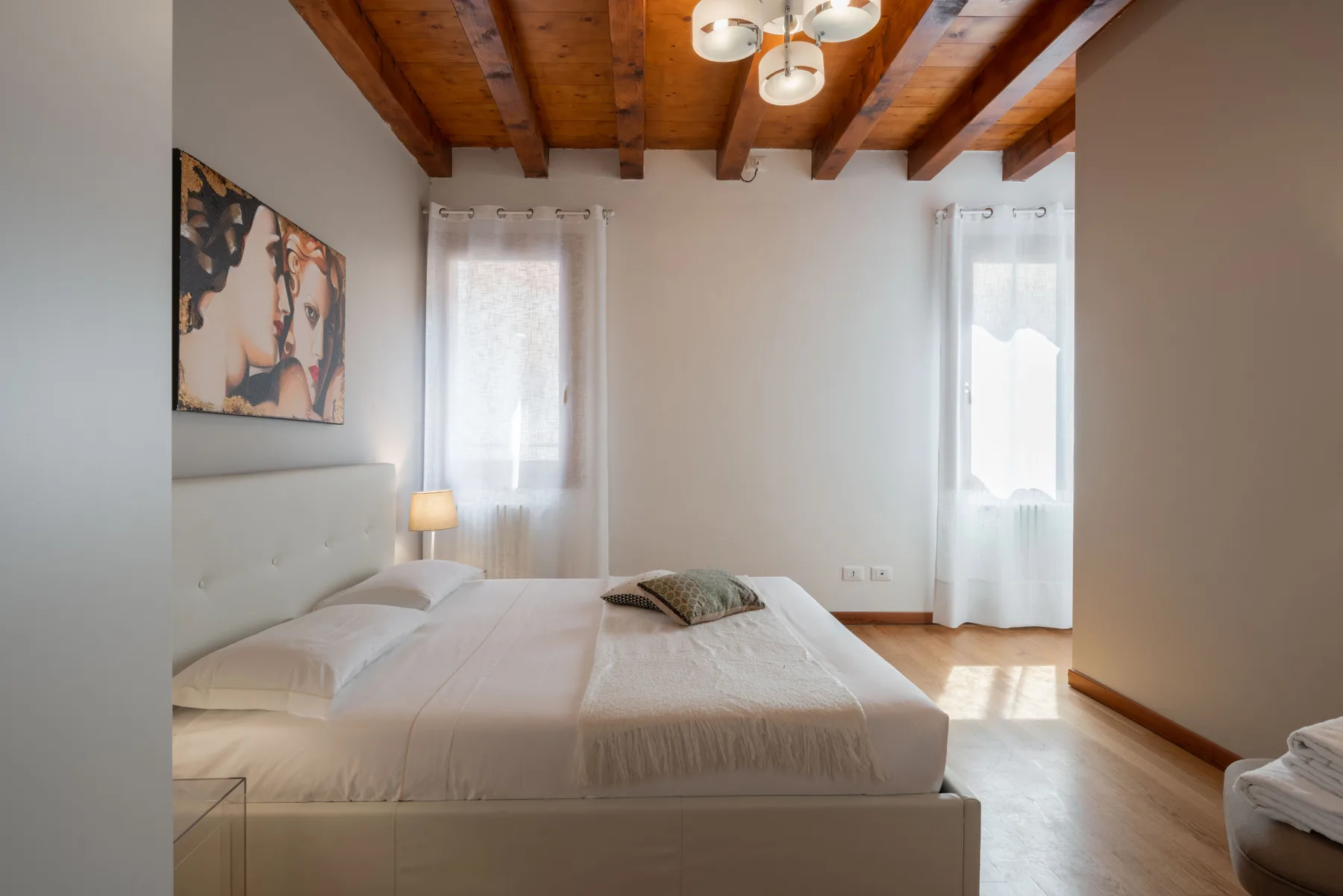 Un dormitorio luminoso con una cama, dos ventanas con cortinas y vigas de madera a la vista.