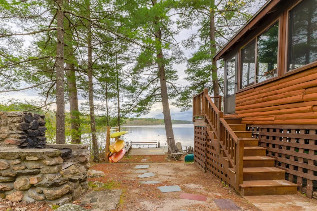 Una cabaña junto a un lago, rodeada de árboles, con una terraza de madera y kayaks cerca de la orilla.