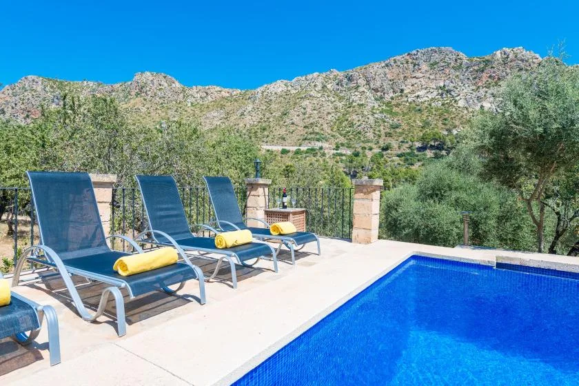 A blue swimming pool and sun loungers sit on a terrace overlooking a rocky hillside.