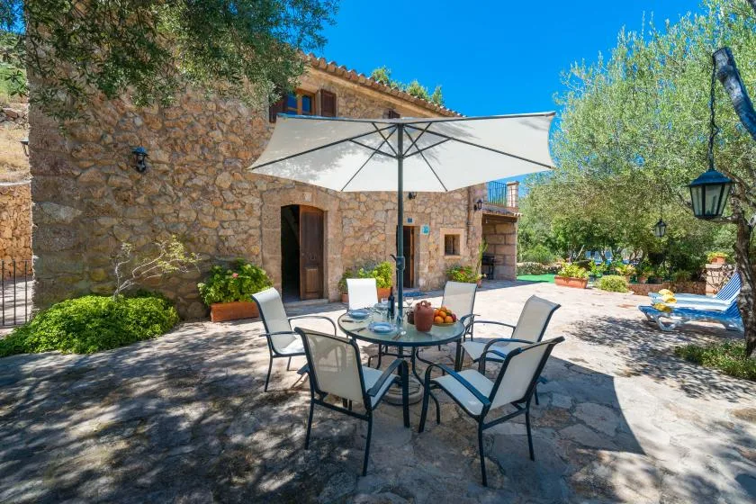 A stone house opens onto a sunny patio with outdoor seating beneath a large umbrella.