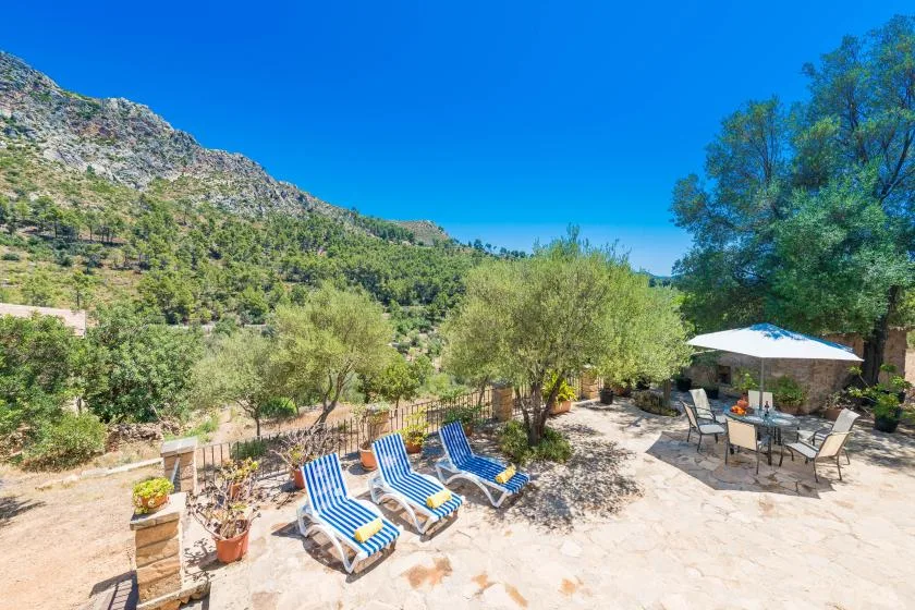 A sunny outdoor patio with lounge seating and an umbrella beside a green hillside.