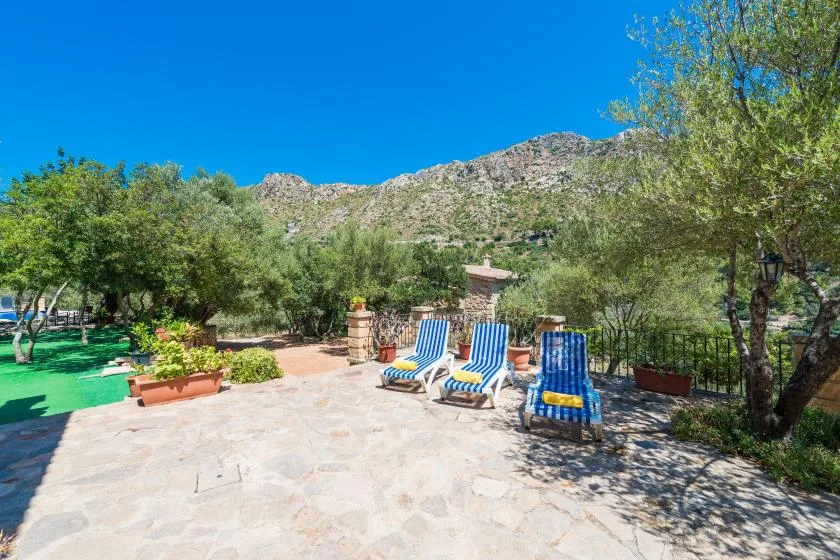 A sunny stone patio with lounge seating overlooks a green garden and rocky hills.