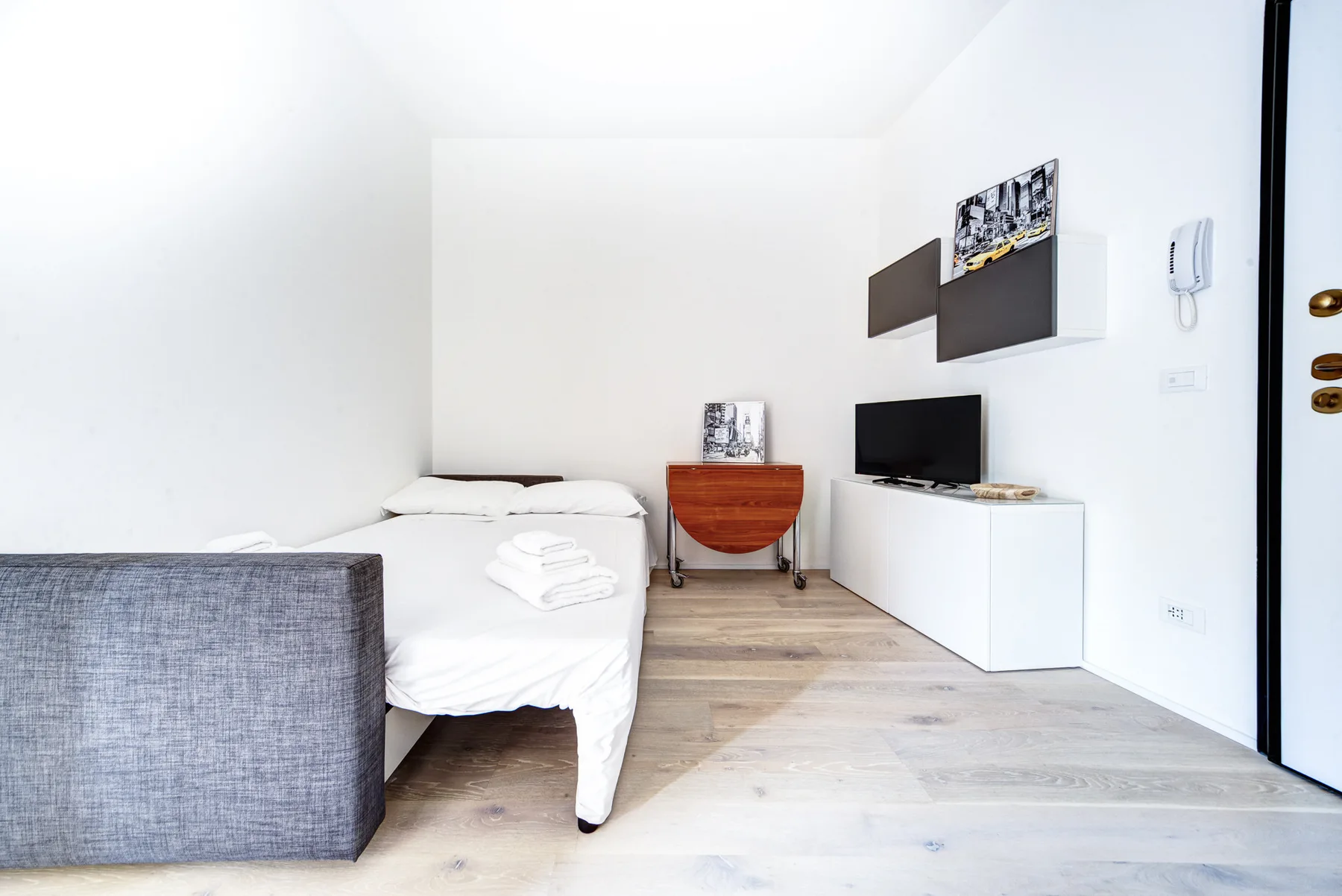 Un dormitorio luminoso con ropa de cama blanca, un televisor y suelo de madera clara.