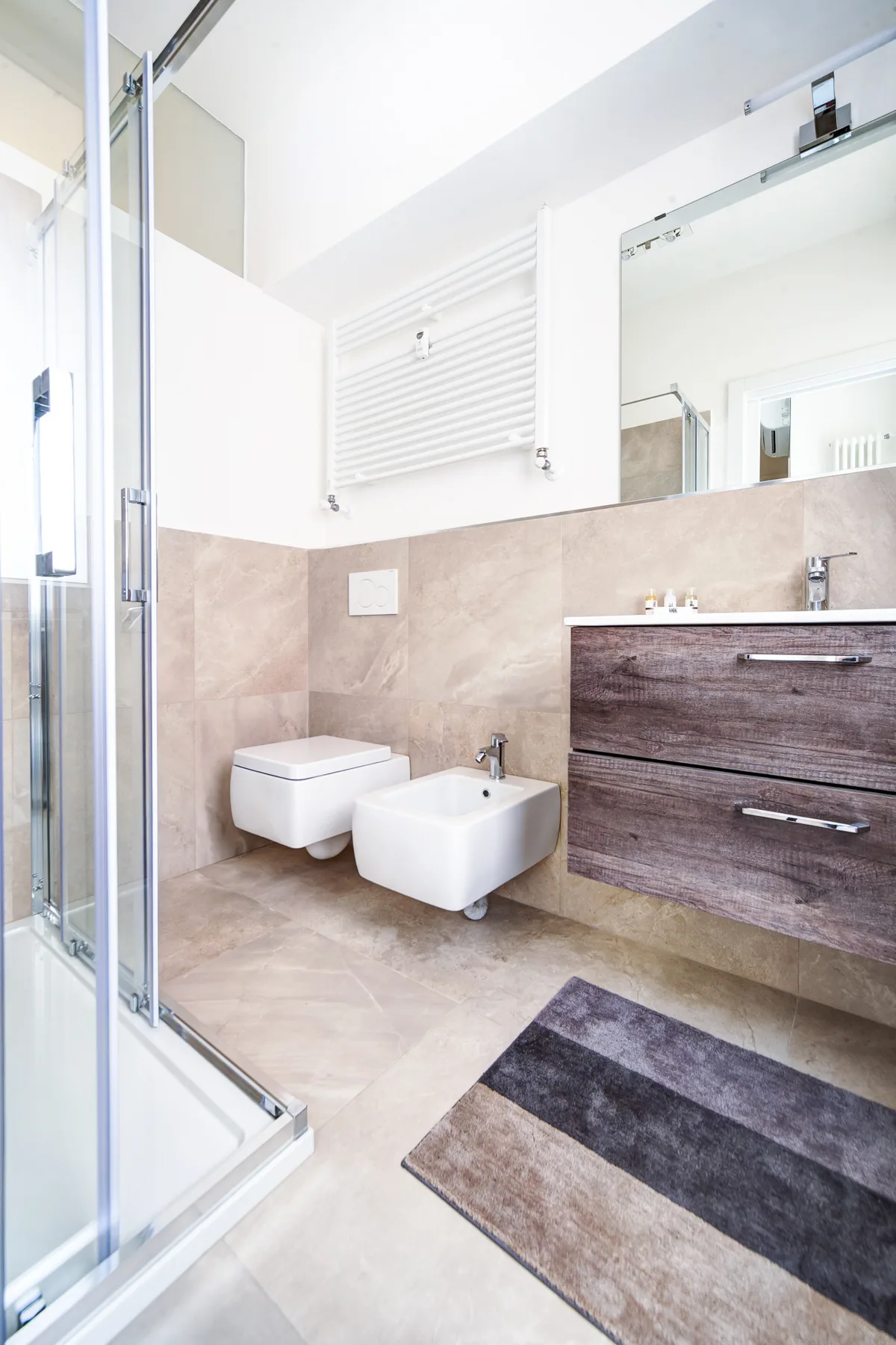 Baño luminoso con ducha de cristal, sanitarios suspendidos y un mueble de madera oscura.
