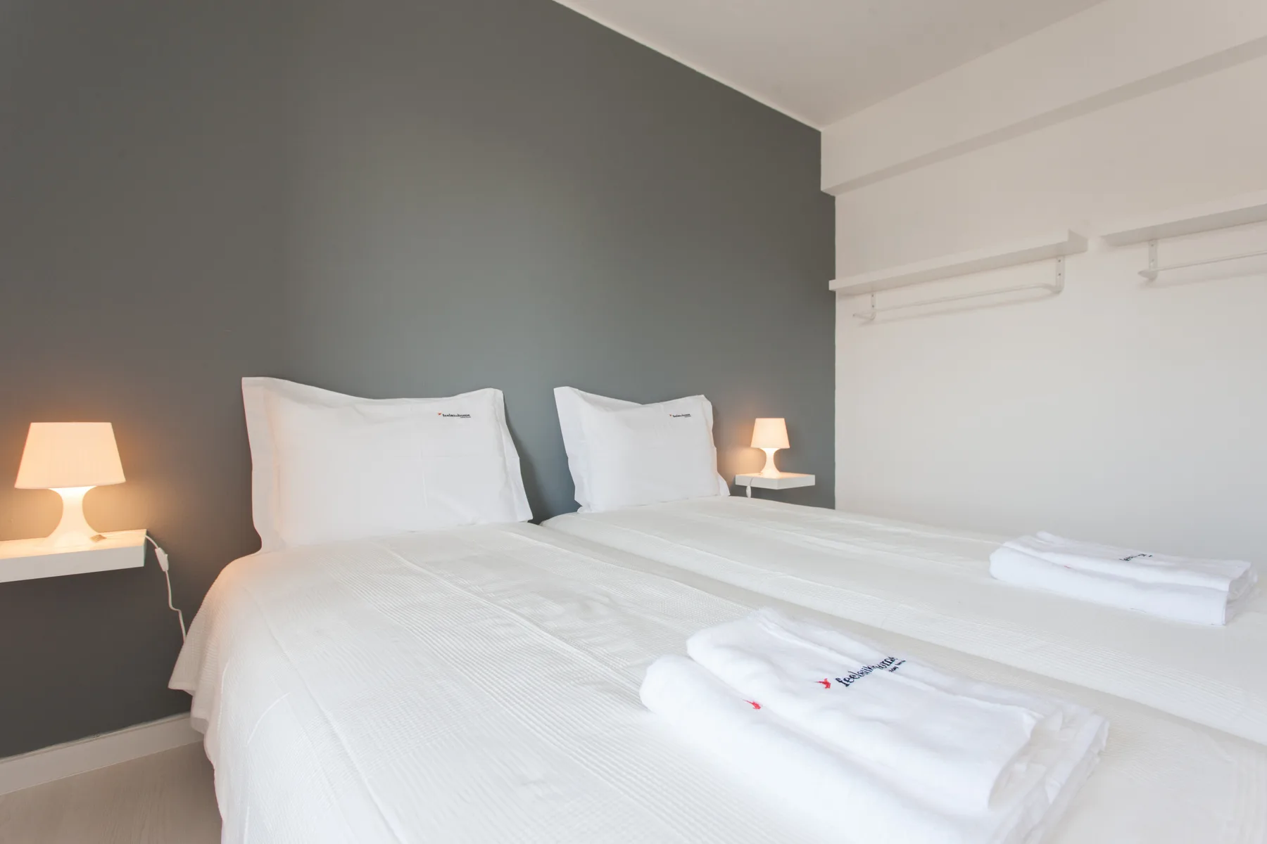 A bedroom with two beds, white linens, and bedside lamps against a gray wall.