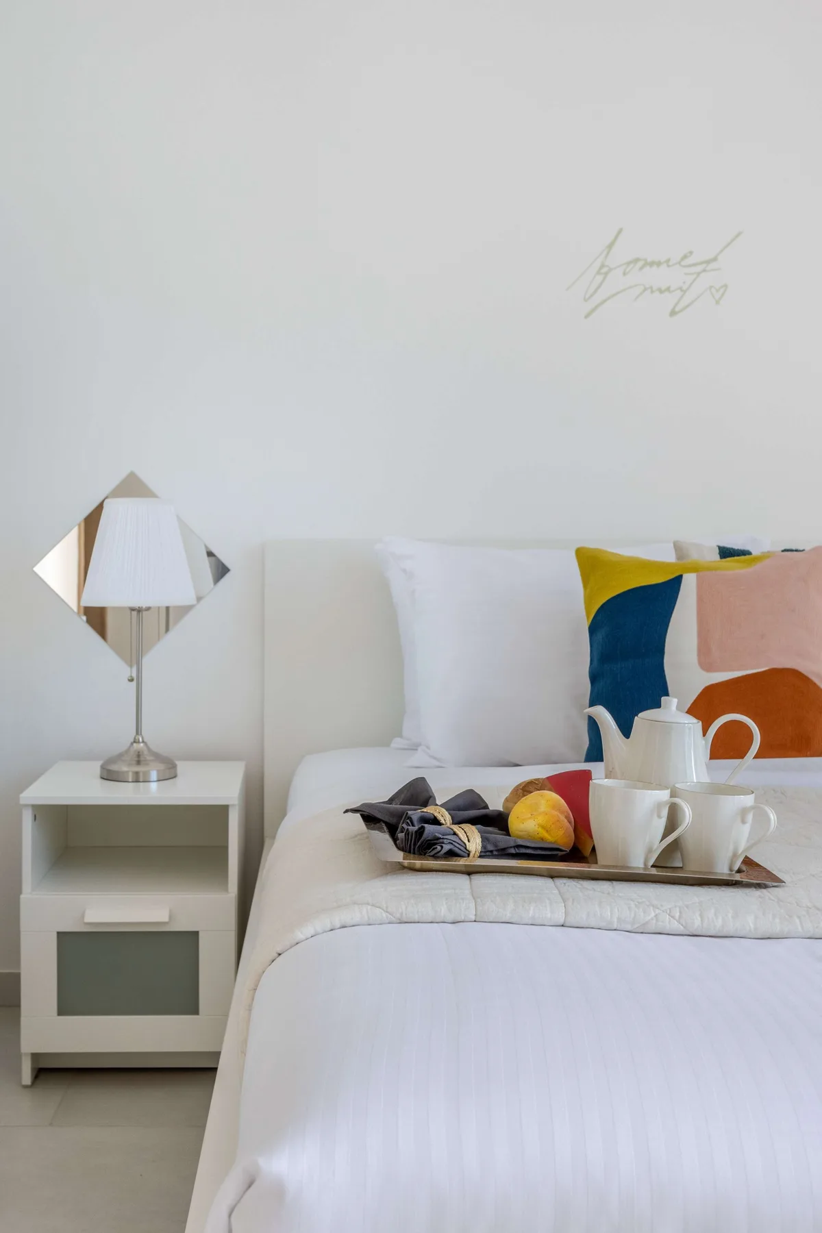 A bedroom with white bedding, colorful accent pillows, and a bedside table.