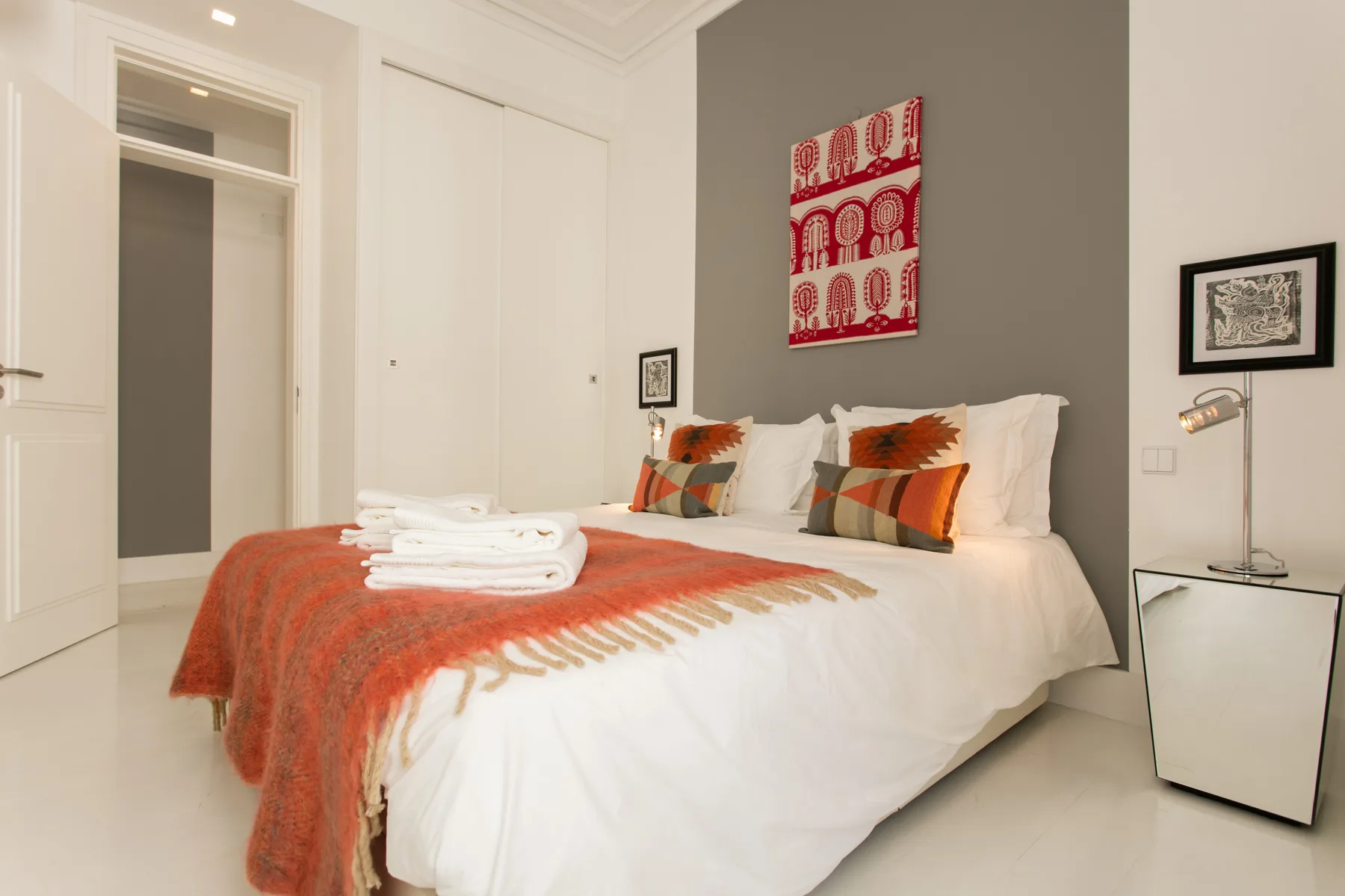 A bright bedroom with a white bed and orange throw against a gray accent wall.