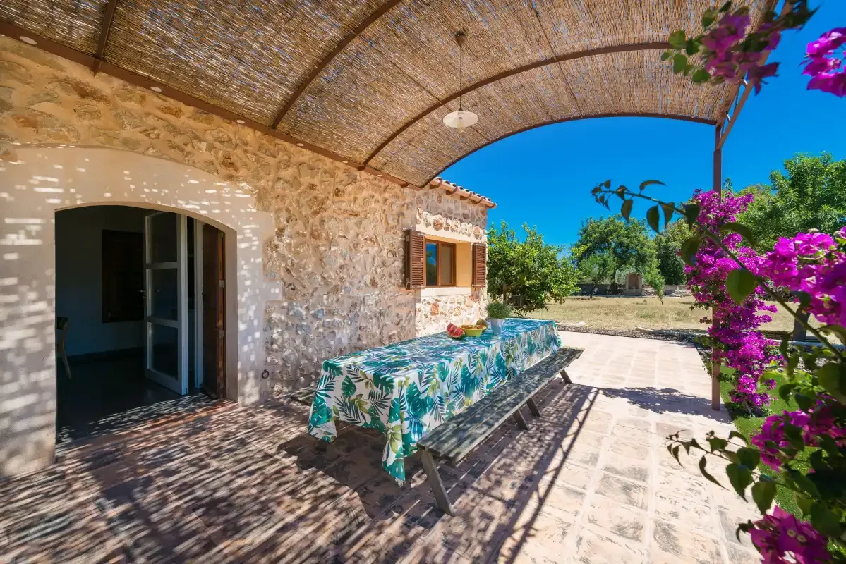 A stone house opens onto a shaded patio with a table and flowering vines.