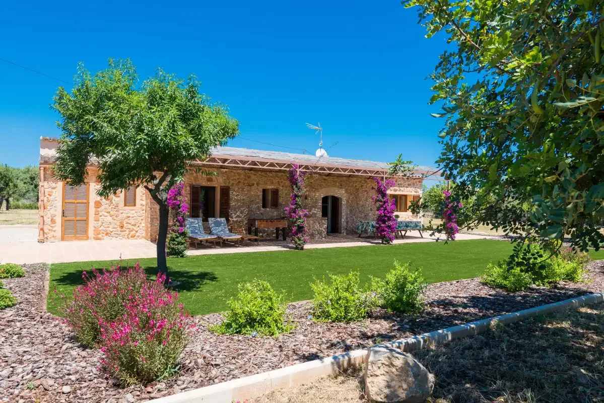 A stone house opens onto a landscaped garden with flowering shrubs and trees.