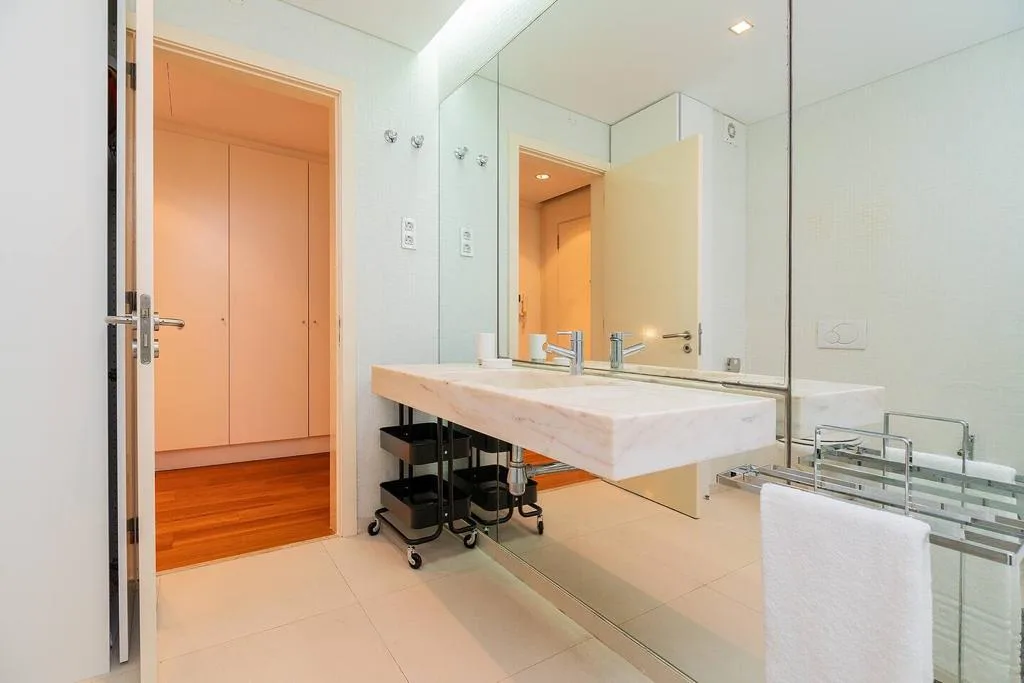Un baño luminoso con un lavabo largo, un espejo de pared y una puerta abierta.